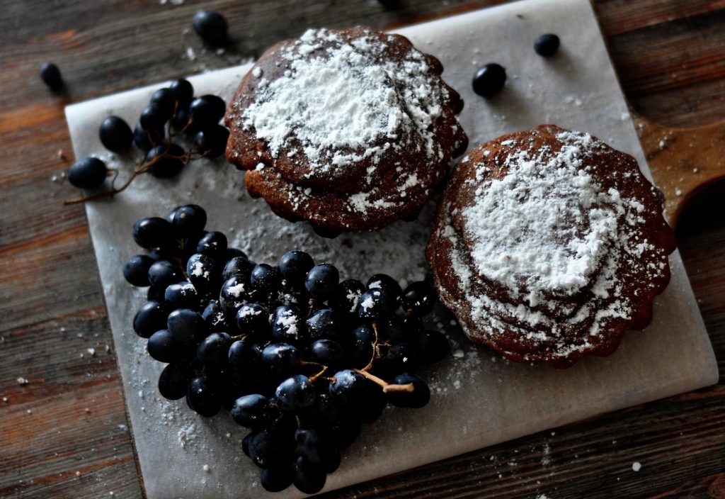 A dessert platter with two cupcakes covered in icing sugar. On the platter are also a bunch of black grapes.
